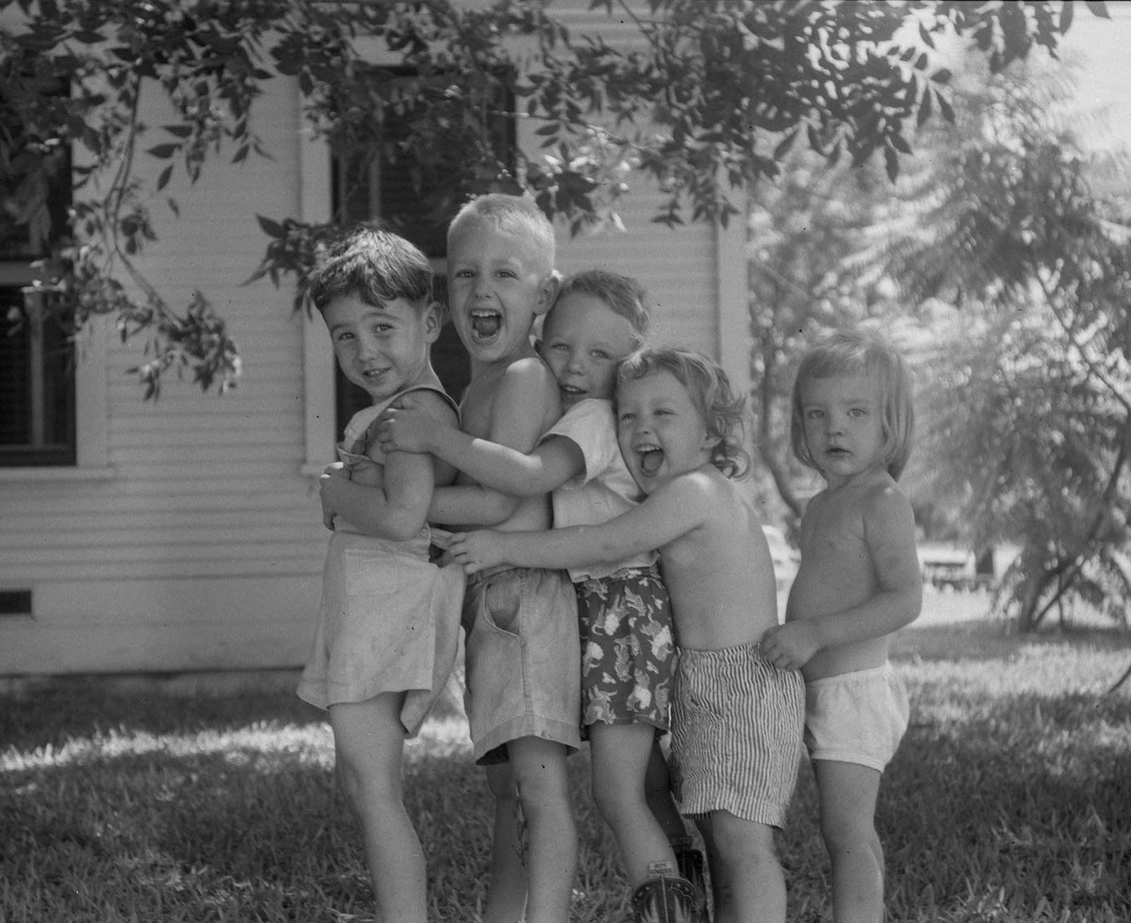 Five young children in a row in a sunlit yard, arms wrapped around one another, most of them laughing