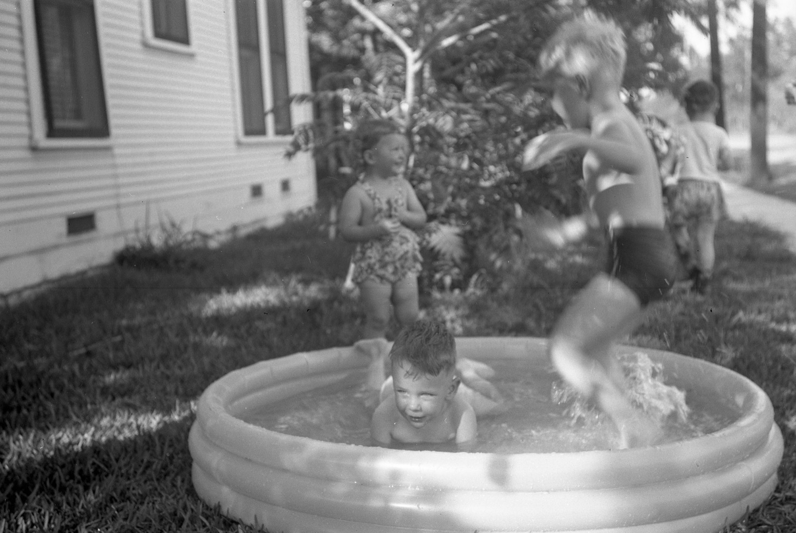 Two boys playing in an inflatable wading pool in a yard, one sitting in the water and one leaping in